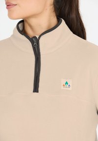 Beige fleece pullover met een zwarte ritssluiting en kraag, voorzien van een klein, gekleurd logo patch. Soepele textuur en een kwart-rits ontwerp.