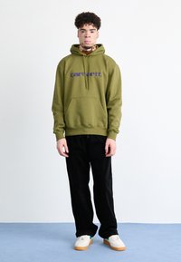 Felpa verde oliva con tasca frontale e logo viola, abbinata a pantaloni in corduroy neri e sneakers bianche con dettagli verdi.