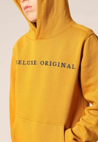 Gele hoodie met de tekst "DELUXE ORIGINAL" in donkerblauwe borduurwerk, een voorzak met kangaroo-stijl en geribbelde manchetten. Zachte textuur.