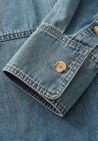 Mansetto di camicia in denim di colore azzurro chiaro, con chiusura a bottone e cuciture arancioni a contrasto lungo i bordi e le cuciture.