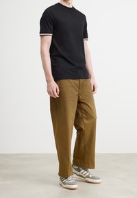 Zwart T-shirt met korte mouwen en witte en bruine gestreepte accenten bij de manchetten, gecombineerd met bruine broek met wijde pijpen en grijze sneakers.