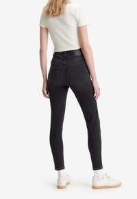 Zwarte high-waisted skinny jeans met een gladde textuur, achterzakken en gestikte details, gecombineerd met een witte geribbelde top met korte mouwen en sneakers.
