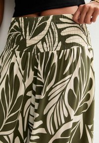 Green and cream-colored trousers with a leafy pattern, featuring a smocked waistband and lightweight fabric.
