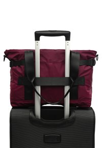 Černý kufr s vínovou polstrovanou taškou zajištěnou nahoře. Taška má černé popruhy a rukojeť, s viditelnými hliníkovými teleskopickými rukojeťmi.