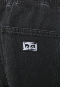 Black denim fabric featuring a visible pocket with a rectangular tag. The tag displays white lettering and a stylized logo with two hands.