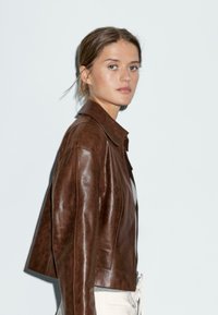 Veste en cuir marron courte avec un col pointu, dotée de manches longues et d'une texture lisse, associée à un pantalon de couleur claire.