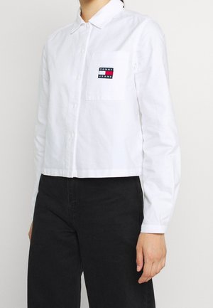 Femme portant une chemise blanche à boutons avec un logo Tommy Jeans sur la poche poitrine et un pantalon noir taille haute sur un fond uni.