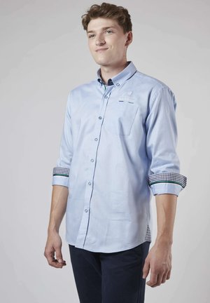 Chemise à boutons bleu clair à manches longues avec un accent à carreaux sur les poignets retroussés ; dotée d'une poche poitrine et d'un logo brodé.