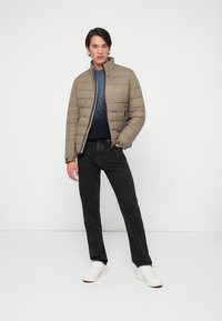 Vadderad olivgrön jacka med hög krage, dragkedjestängning och tonal logotyp, ihop med mörka jeans och vita sneakers, står mot en vit bakgrund.