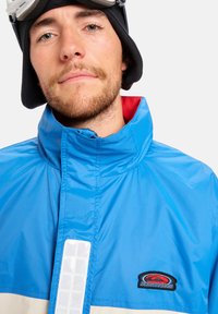 Μπλε αδιάβροχη ζακέτα με ψηλό γιακά, διαθέτει κόκκινη εσωτερική επένδυση και κεντημένο λογότυπο. Στο μπροστινό μέρος υπάρχει διαφανής πλαστική τσέπη για λεπτομέρειες.