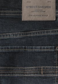 Ciemnoniebieskie dżinsy z skórzaną metką z wytłoczonym tekstem. Wyraźne przeszycia w kontrastowym beżowym kolorze podkreślają design.