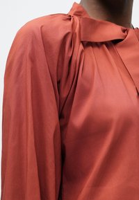 Blusa color ruggine con collo alto, design delle spalle arricciato e tessuto leggero e morbido. Presenta dettagli minimalisti.