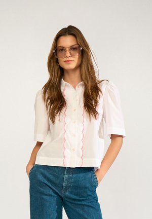 Kobieta nosząca duże okulary przeciwsłoneczne, białą bluzkę z krótkim rękawem z różowym falistym obszyciem i niebieskie dżinsy, stojąca z rękami w kieszeniach.