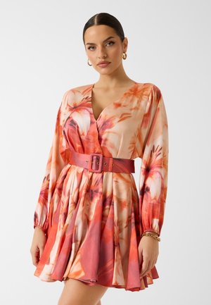 Kobieta w długiej sukience w kwiaty, pomarańczowo-różowej, z szerokim dopasowanym paskiem i złotymi kolczykami kołami, stojąca na białym tle.