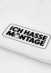 White knitted beanie with a textured surface and a rectangular black and white patch featuring the text "¡CH HASSE MONTAGE!" and a penguin graphic.