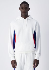 Sweatshirt à capuche blanche avec des accents latéraux bleus et rouges, fabriqué dans un matériau doux et avec une coupe décontractée. Dispose d'une poche kangourou et de poignets côtelés.