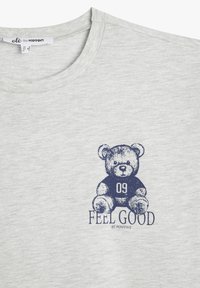 Camiseta de algodón gris con un oso de peluche impreso en azul, con el número de jugador 09 y el texto "SIENTE BIEN, SÉ POSITIVO" debajo.