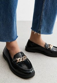 I mocassini in vernice nera presentano un accentuato con una catena dorata, con punta arrotondata e dettagli testurizzati. Abbinati a jeans in denim blu a vita bassa.