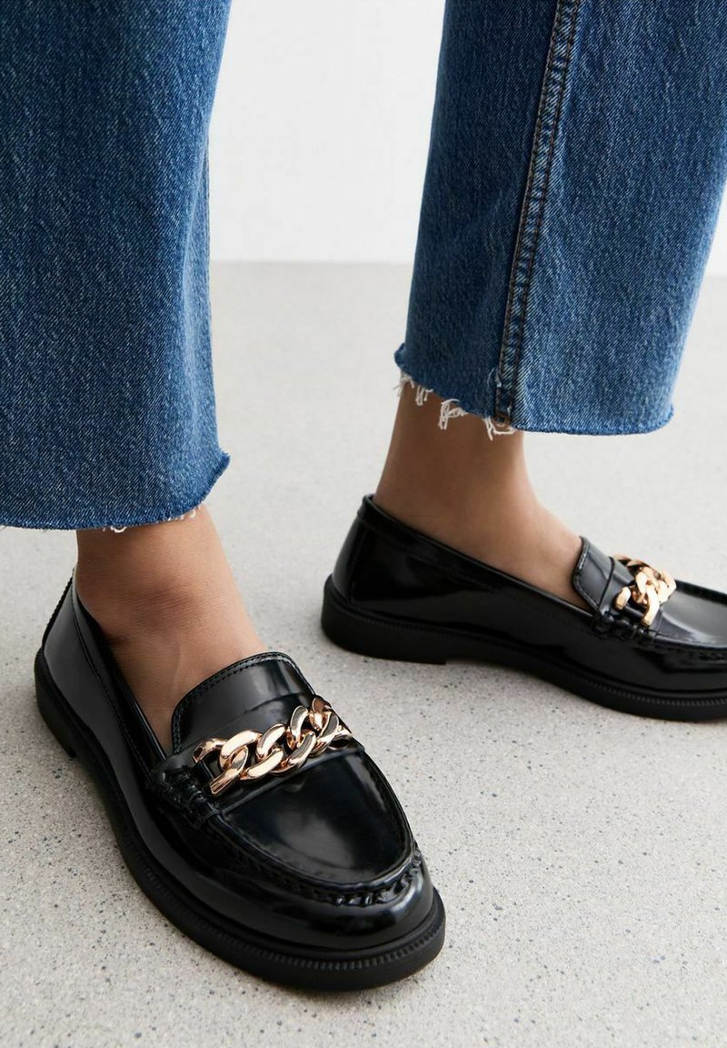 I mocassini in vernice nera presentano un accentuato con una catena dorata, con punta arrotondata e dettagli testurizzati. Abbinati a jeans in denim blu a vita bassa.