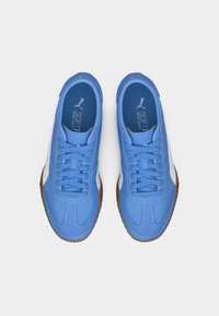 Paire de baskets Puma bleues avec des rayures blanches, lacets noués, et semelles en caoutchouc marron, vues de dessus sur un fond blanc.