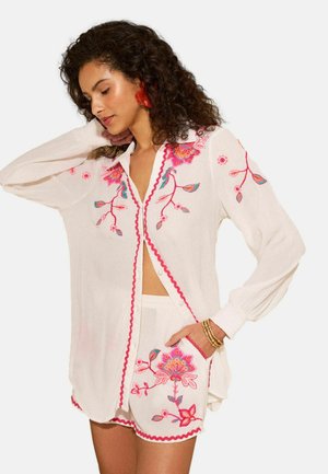 Žena oblečená v bielom košeľovom tričku na gombíky a šortkách s ružovo-červenou kvetinovou výšivkou pózuje s jednou rukou za hlavou.