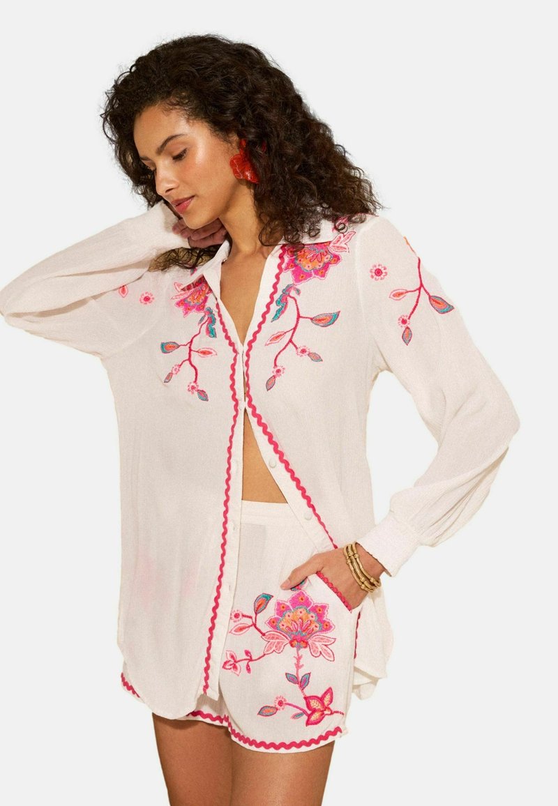 Frau trägt ein weißes Hemd mit Knöpfen und Shorts mit rosa und roter Blumenstickerei, posiert mit einer Hand hinter dem Kopf.