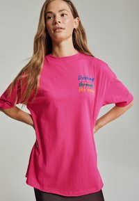 Rosa T-Shirt mit dem Text "Durch den Schnee flitzen" in bunten Buchstaben. Kurze Ärmel, lockere Passform und weicher Stoff.