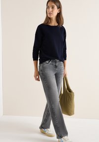 Maglione a maglia blu navy, jeans grigi e sneakers bianche con dettagli blu. Tiene una borsa tote verde. In piedi contro uno sfondo neutro.