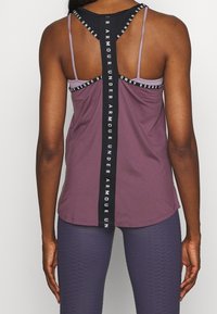 Lila atletisk ärmlös topp med tunna band, svart logotypdetalj på ryggen, slät textur och rundad fåll. Matchad med texturerade leggings.