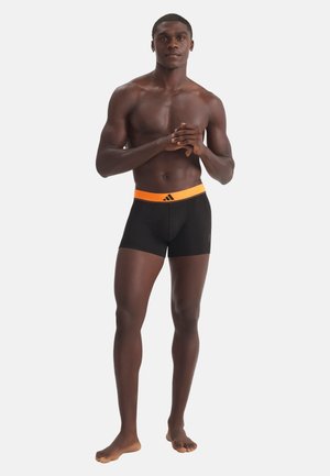 Athletischer Mann steht barfuß in schwarzen Boxershorts mit orangefarbenem Bund vor einem schlichten hellen Hintergrund.