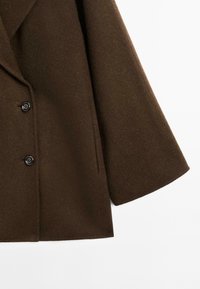 Bruine wolmix jas met een dubbele rij knopen, zwarte knopen en een licht uitlopende mouw. Glad textuur en minimale details.
