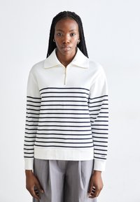 Felpa bianca con righe orizzontali blu navy, colletto a zip a un quarto, polsini e orlo a coste. Abbinata a pantaloni grigi a gamba larga.