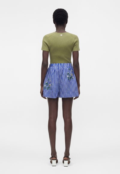 Femme debout de dos, portant un top vert à manches courtes, un short rayé bleu et blanc avec broderie florale, et des sandales noires à talons.