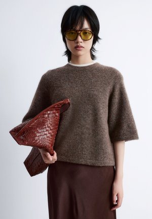 Brown fuzzy sweater with short sleeves, worn over a brown satin skirt; holding a woven red handbag with a triangular shape.