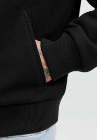 Zwarte sweatshirt met een gestructureerde stof, ribbelboorden en zoom, met een hand in een zijzak gestoken.