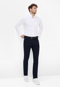 Camisa blanca de botones con mangas largas, pantalones azul marino y zapatillas blancas. El conjunto presenta una silueta ajustada y un diseño clásico.