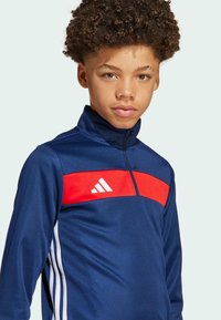 Marineblauwe sportieve zip-up jack met een rode horizontale streep en wit Adidas-logo. Bevat een hoge kraag en witte zijstrepen. Zachte textuur.
