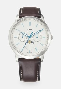 Montre en acier inoxydable argenté avec un cadran blanc, des aiguilles bleues et un bracelet en cuir marron. Présente des indicateurs de date et de jour, ainsi qu'un détail sur la phase de la lune.
