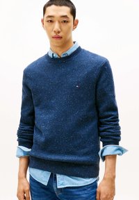 Marineblauer Pullover mit Rundhalsausschnitt, leicht gesprenkelt, Strickstruktur und gerippten Bündchen, getragen über einem hellblauen Hemd mit Kragen.