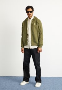 Sweatshirt verde-azeitona com capuz, logotipo dourado no peito, combinado com uma camisa branca, calças pretas e sapatilhas brancas num fundo de carpete azul.