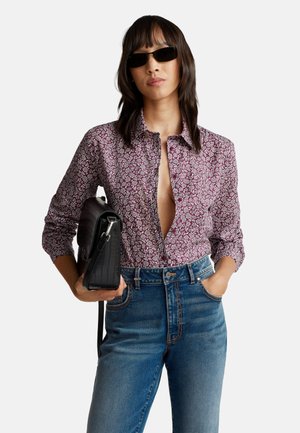 Donna con i capelli lunghi fino alle spalle, che indossa occhiali da sole neri, una camicia button-down floreale bordeaux, jeans blu e tiene una borsa nera sotto il braccio.