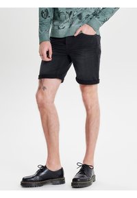 Persona che indossa pantaloni corti neri arrotolati, una camicia a maniche lunghe verde con stampe botaniche e scarpe nere stringate, in piedi con una mano in tasca.