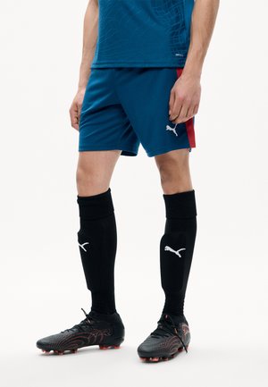 Hombre con pantalones cortos de fútbol Puma azules con una raya roja, calcetines Puma negros hasta la rodilla y botas negras con tacos rojos, de pie sobre fondo blanco.