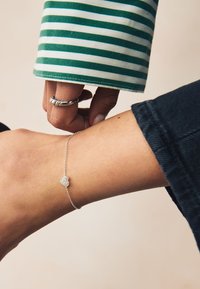 Silbernes Armband mit einem herzförmigen Anhänger, verziert mit kleinen klaren Kristallen. Getragen an einem Handgelenk mit einer dunklen Jeansärmel und grün gestreiftem Stoff.