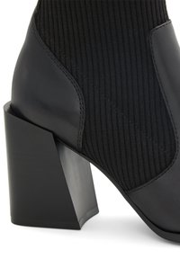 Bottine noire avec un dessus en tissu texturé et des accents en cuir lisse, présentant un talon géométrique épais et des détails de couture minimalistes.