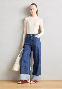 Haut à manches longues en ribbed crème avec un col à volants, jean bleu taille haute à jambes larges avec des poignets en denim contrasté, et des sandales rouges à clous.
