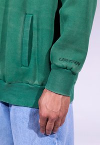 Felpa verde con orlo e polsini a coste, tasca laterale e logo "URTRN" ricamato sulla manica. Abbinata a jeans azzurri.