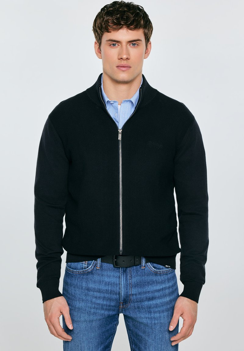 Fekete cipzáras pulóver bordázott textúrával és magas gallérral, világoskék galléros inggel és kék farmernadrággal párosítva.