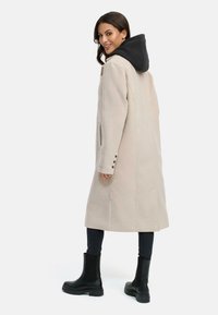 Beige lange jas met een zwarte capuchon, aangesloten zijzakken en knoopaccenten. Het model draagt zwarte enkellaarzen en donkere jeans.