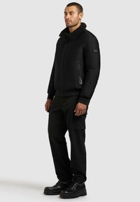 Svart bomberjakke med en fleecekrage, glidelåslommer og elastiske mansjetter, kombinert med svarte cargobukser og svarte snøresko.